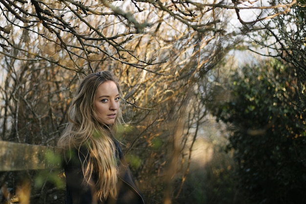 Young blonde female with a black coat standing on pathway surrounded by leafless trees