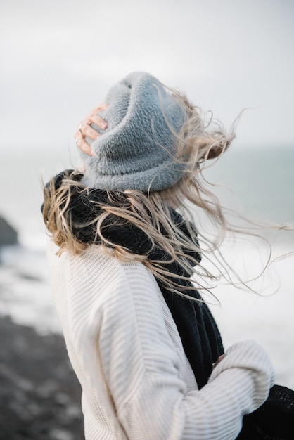 Молодая блондинка в шляпе гуляет по скалистому морскому берегу в ветреную погоду
