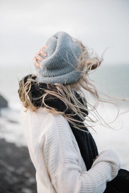 Young blonde female with a hat walking on a rocky sea coast in a windy weather