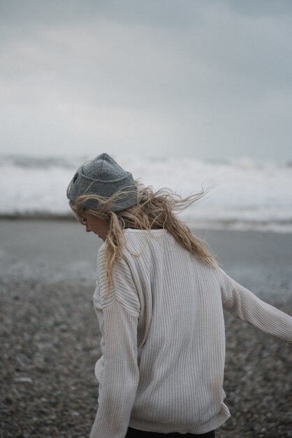 Young blonde female with a hat walking on a rocky sea coast in a windy weather