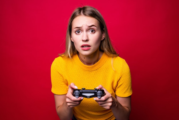 Бесплатное фото Молодая блондинка-геймер с помощью геймпада играет в видеоигры, изолированные на красной стене