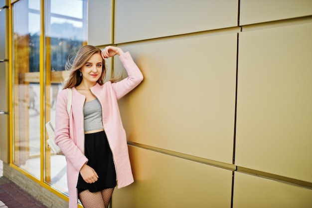 Free photo young blonde girl in black skirt and pink coat posed against golden texture wall