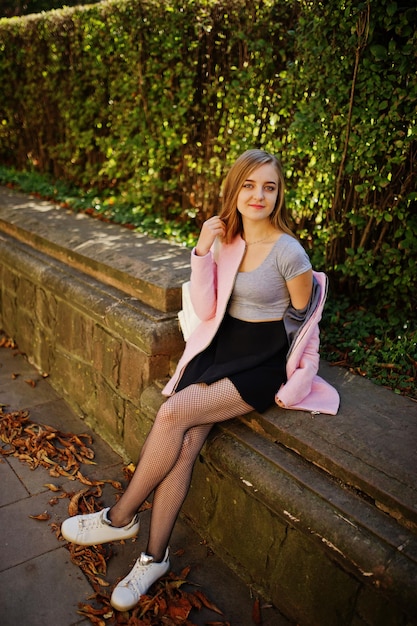 Молодая блондинка в черной юбке и розовом пальто позирует в солнечный день на улице
