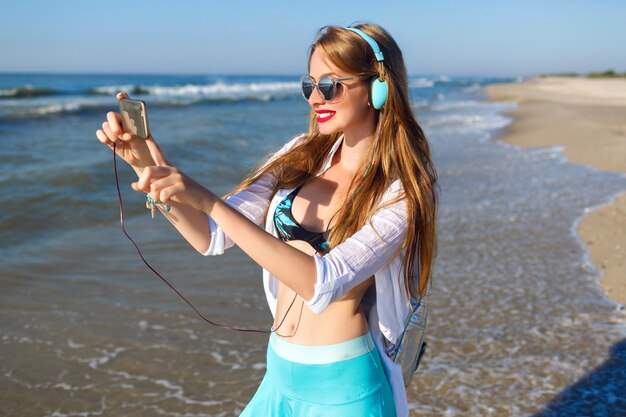 Молодая блондинка веселится на пляже, яркое закрытие битника, отдых у океана, слушает расслабляющую музыку и делает селфи на своем телефоне.