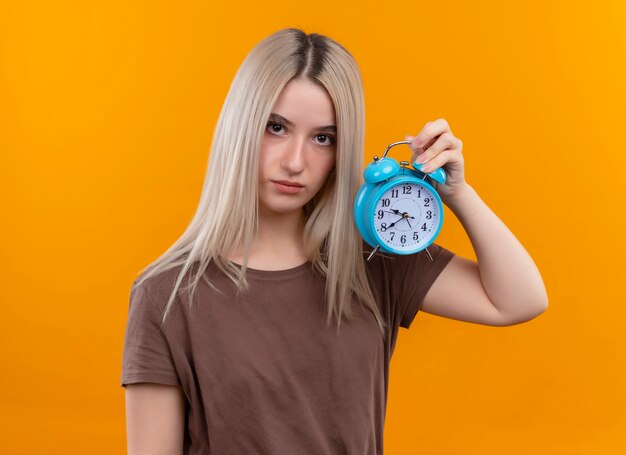 Молодая блондинка держит будильник на изолированной оранжевой стене