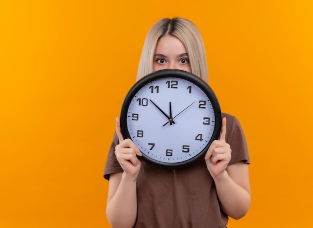 Young blonde girl holding clock hiding behind it on isolated orange wall with copy space