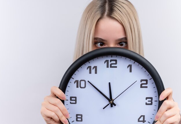 Young blonde girl holding clock and hiding behind it with crossed eyes on isolated white wall with copy space