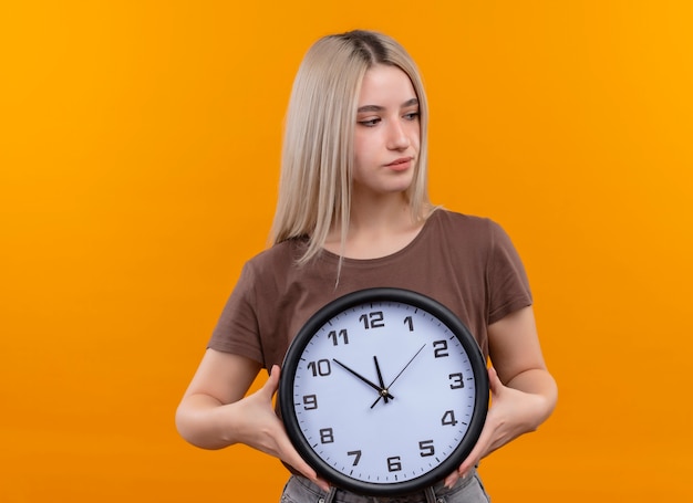Young blonde girl holding clock looking at right side on isolated orange wall with copy space
