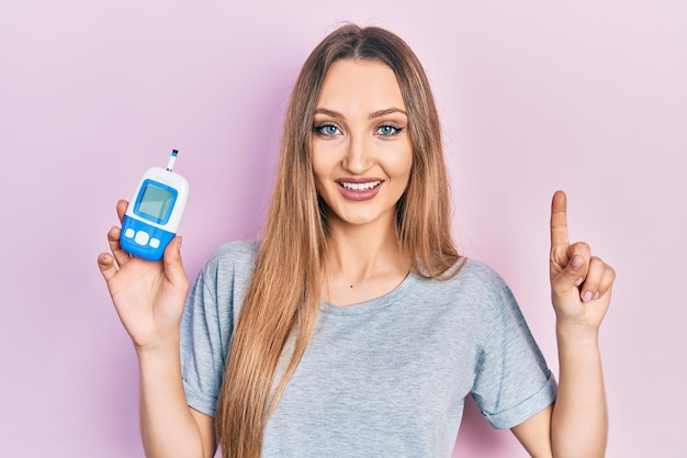 Free photo young blonde girl holding glucometer device smiling with an idea or question pointing finger with happy face number one