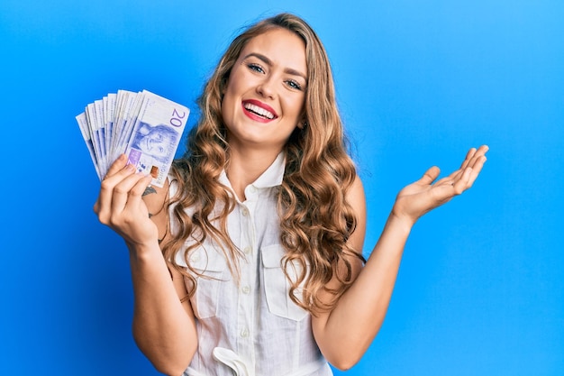 Young blonde girl holding swedish krona banknotes celebrating achievement with happy smile and winner expression with raised hand