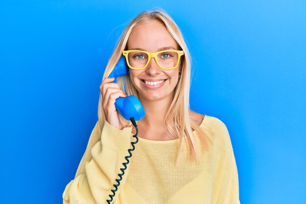 Молодая блондинка, держащая старинный телефон, выглядит позитивно и счастливо, стоя и улыбаясь с уверенной улыбкой, показывая зубы
