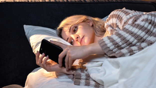 Young blonde girl is on her smartphone in the bed. Trying to fall asleep. Social media addiction