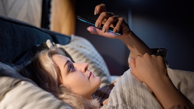 若いブロンドの女の子は、ベッドの中で彼女のスマートフォンにいます。眠ろうとしています。ソーシャルメディア中毒