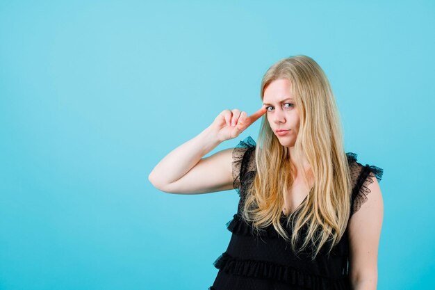 Young blonde girl is looking at camera by holding forefinger on temple on blue background