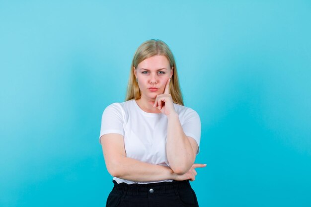 Young blonde girl is looking at camera by putting forefinger on cheek on blue background