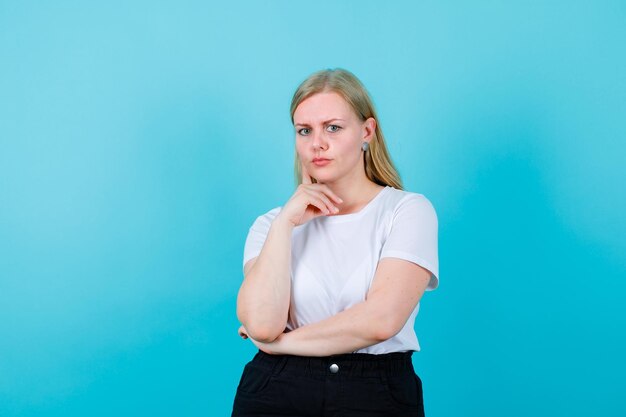 Young blonde girl is looking at camera by putting forefinger on cheek on blue background