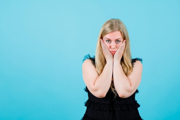 Young blonde girl is looking at camera by putting hands on cheeks on blue background