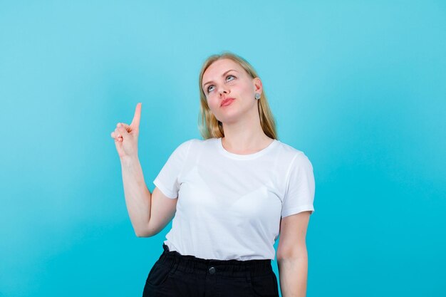 Young blonde girl is looking and pointing up with forefingers on blue background