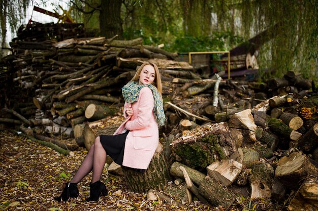 나무 그루터기 배경에 포즈를 취한 분홍색 코트를 입은 젊은 금발 소녀