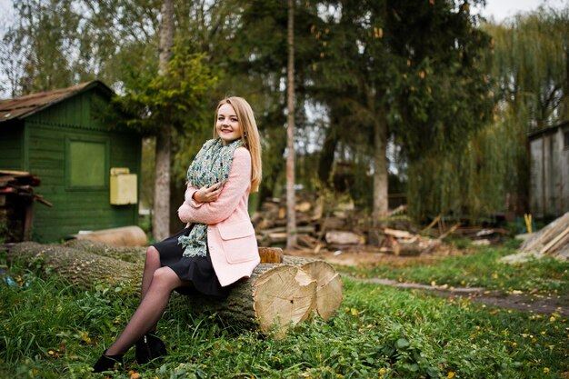 Young blonde girl at pink coat posed on autumn park sitting on cut tree against house of the forester