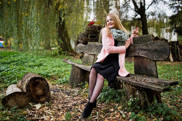 Молодая блондинка в розовом пальто сидит на скамейке на фоне деревянных пней
