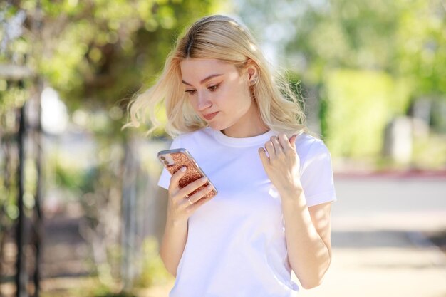 Молодая блондинка стоит и смотрит на свой телефон в парке Фото высокого качества