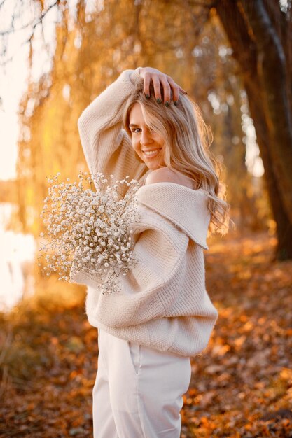 Free photo young blonde girl with flowers standing in autumn park near the lake woman wearing beige sweater girl posing for a photo in sunny day