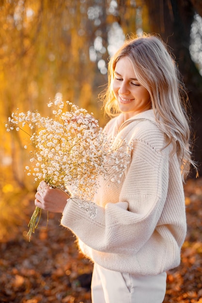 Молодая блондинка с цветами стоит в осеннем парке у озера Женщина в бежевом свитере и белых брюках Девушка позирует для фото в солнечный день
