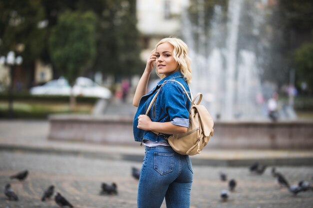 Young blonde girl woman on streetwalk fontain dressed up in blue jeans suite with bag on her shoulder in sunny day