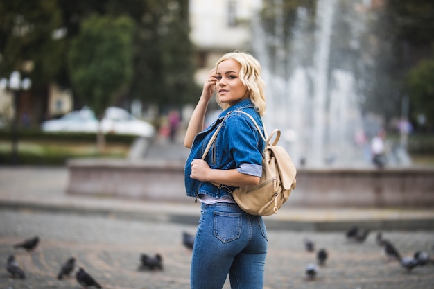 Молодая блондинка женщина на уличном фонтане, одетая в синие джинсы с сумкой на плече в солнечный день