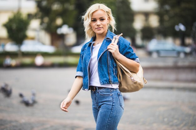 Young blonde girl woman on streetwalk square fontain dressed up in blue jeans suite with bag on her shoulder in sunny day