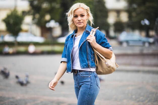 Молодая блондинка женщина на уличном квадратном фонтане, одетая в синие джинсы с сумкой на плече в солнечный день