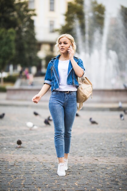 Young blonde girl woman talks on phone on streetwalk square fontain dressed up in blue jeans suite with bag on her shoulder in sunny day