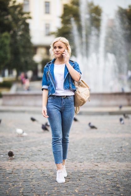 Молодая блондинка разговаривает по телефону на уличном квадратном фонтане, одетая в синие джинсы с сумкой на плече в солнечный день