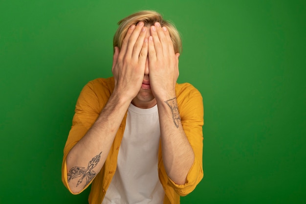 Young blonde guy wearing yellow t-shirt covered face with hands isolated on green with copy space