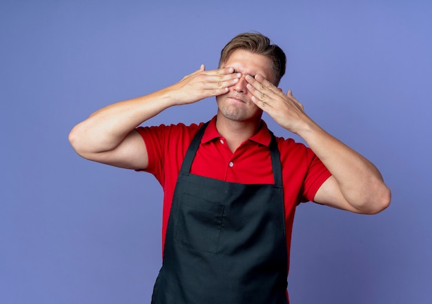 Foto gratuita giovane maschio biondo barbiere in uniforme chiude gli occhi con le mani isolate su sfondo viola