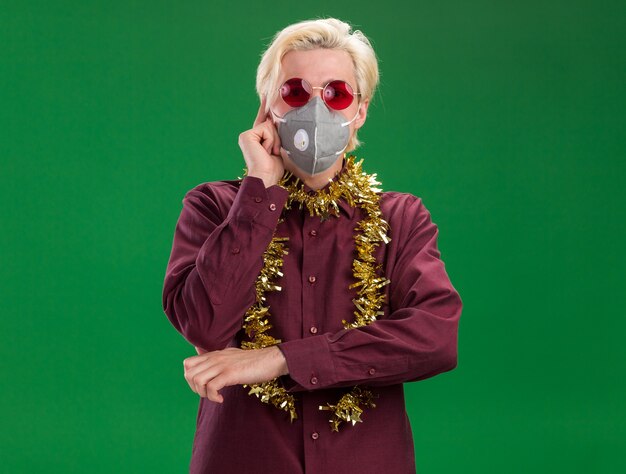 Young blonde man wearing glasses and protective mask with tinsel garland around neck looking at camera doing think gesture isolated on green background with copy space