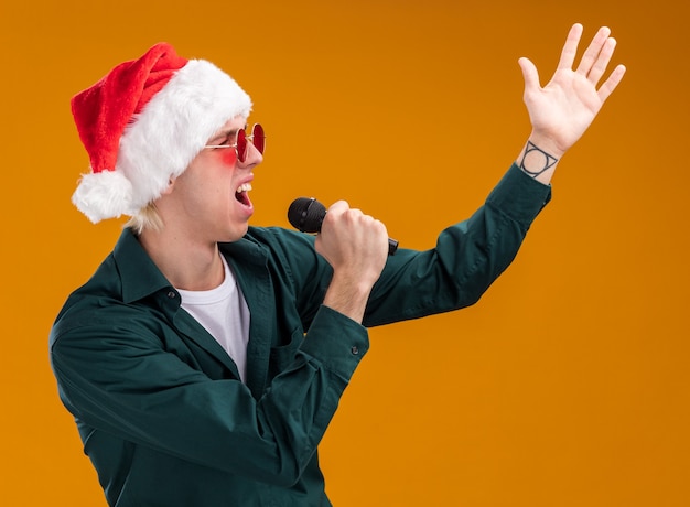 Young blonde man wearing santa hat and glasses holding microphone looking at side keeping hand in air singing isolated on orange background