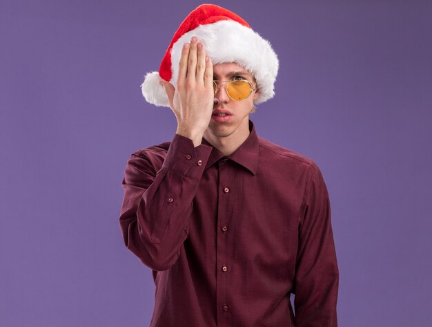 Young blonde man wearing santa hat and glasses looking at camera covering half of face with hand isolated on purple background with copy space