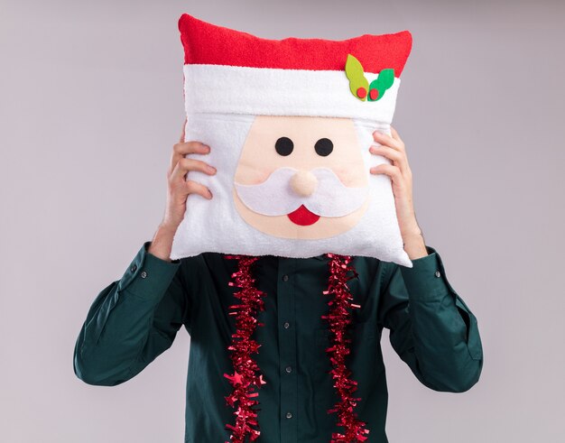 Free photo young blonde man wearing santa hat and glasses with tinsel garland around neck holding santa claus pillow covering face with it isolated on white background