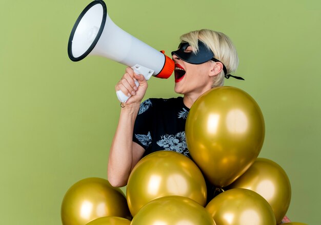 Young blonde party girl wearing masquerade mask standing behind balloons turning head to side talking by speaker with closed eyes isolated on olive green background with copy space