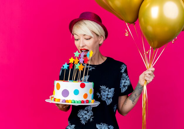 Young blonde party girl wearing party hat holding balloons and birthday cake with stars trying to bite cake isolated on crimson background with copy space