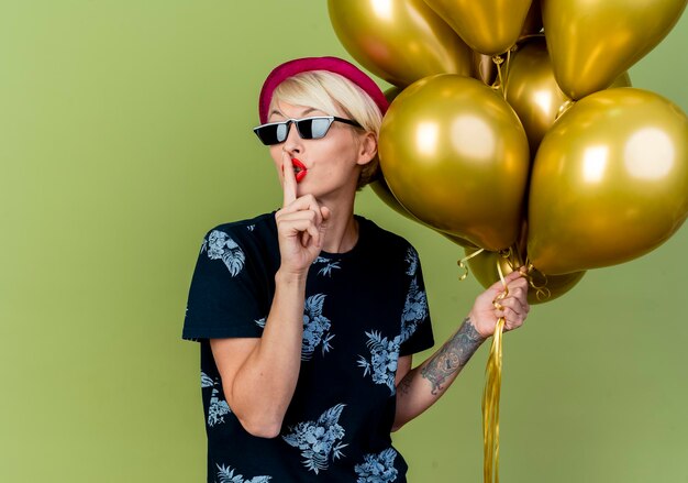 Young blonde party woman wearing party hat and sunglasses holding balloons doing silence gesture isolated on olive green wall with copy space