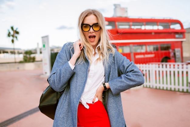 세련된 스마트 캐주얼 학생 복장, 블루 코트 및 색안경, 가을 봄 중반 시즌 시간, 여행 분위기를 입고 런던 도심에서 산책하는 젊은 금발의 예쁜 여자.