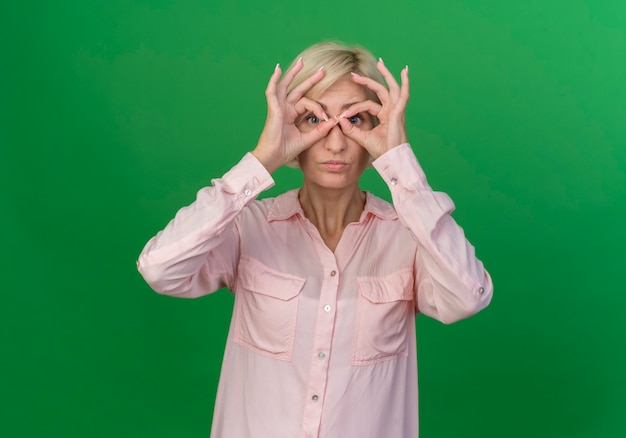 Giovane donna slava bionda che fa gesto di sguardo alla macchina fotografica usando le mani come binocolo isolato su priorità bassa verde