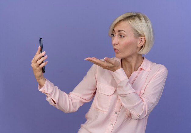 Young blonde slavic woman holding mobile phone and sending blow kiss at it isolated on purple background