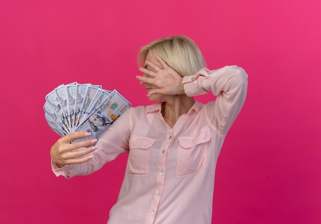 Free photo young blonde slavic woman stretching out money towards camera and hiding face behind hand isolated on pink background with copy space