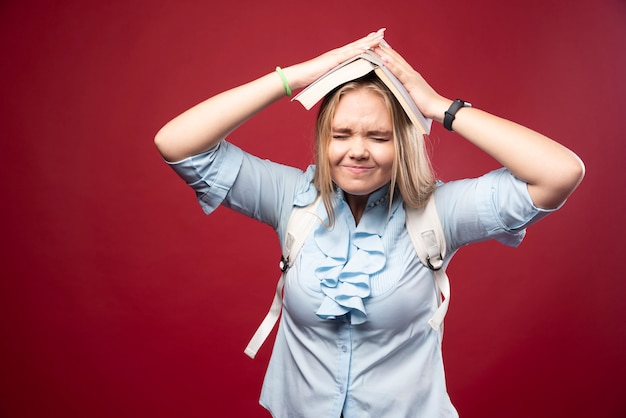 Young blonde student woman holds her book at her head and looks tired and confused.