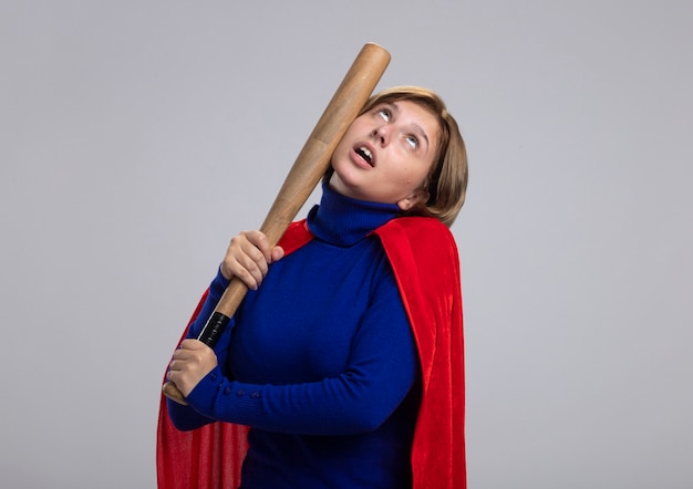 Free photo young blonde superhero girl in red cape beating herself in head with baseball bat looking up isolated on white background with copy space