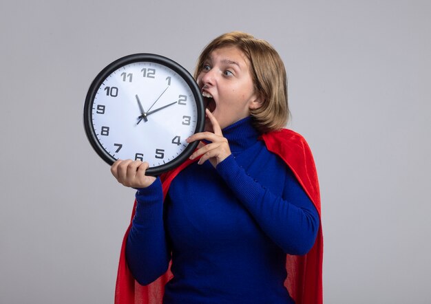 Young blonde superhero girl in red cape holding and looking at clock trying to bite it isolated on white background with copy space